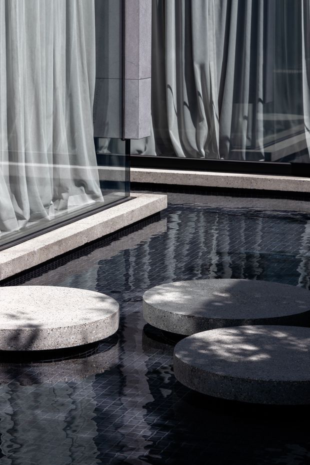 A close-up architectural view of a dark tiled reflecting pool or shallow water feature beside floor-to-ceiling glazing. Three large circular concrete stepping platforms sit in the water, while sheer curtains and reflective glass define the surrounding modern facade. Strong geometric forms, muted grey tones, and dappled sunlight create a calm, refined atmosphere.