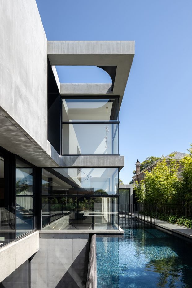 A striking contemporary multi-level residence with exposed concrete slabs, black-framed floor-to-ceiling glazing, and cantilevered architectural volumes arranged beside a narrow blue swimming pool. The view emphasizes the sculptural facade, open-air voids, transparent living spaces, and strong geometric shadows, with mature greenery and neighbouring traditional housing visible beyond.