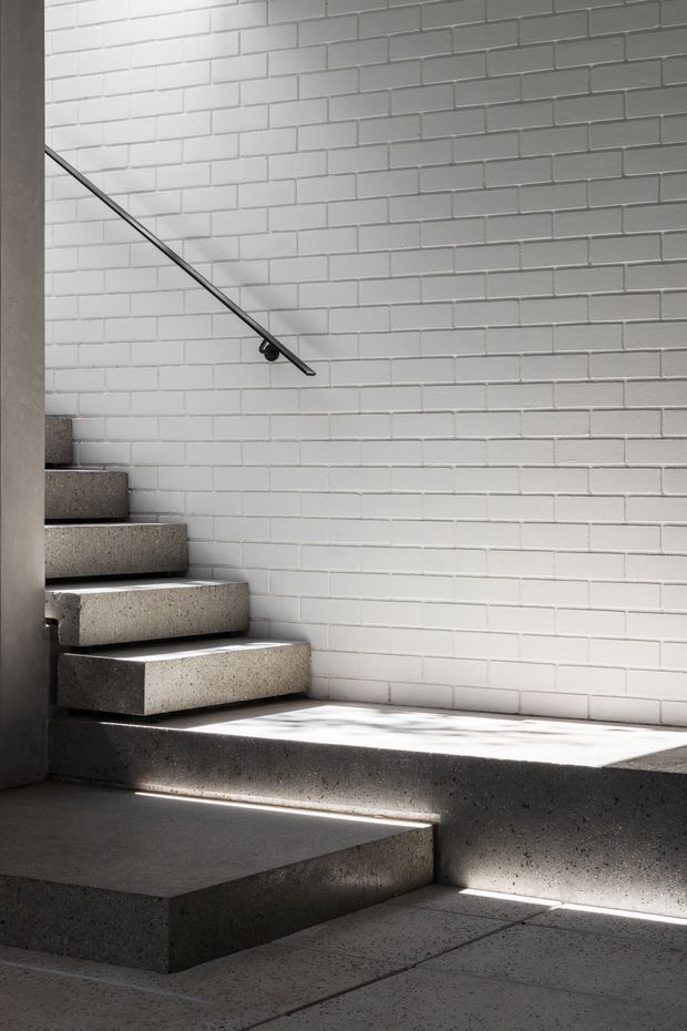 Minimalist interior stairway with floating, solid concrete steps and broad concrete landings. A white painted brick wall forms the backdrop, with a slim dark metal handrail and strong sunlight creating sharp shadows across the monochromatic space.