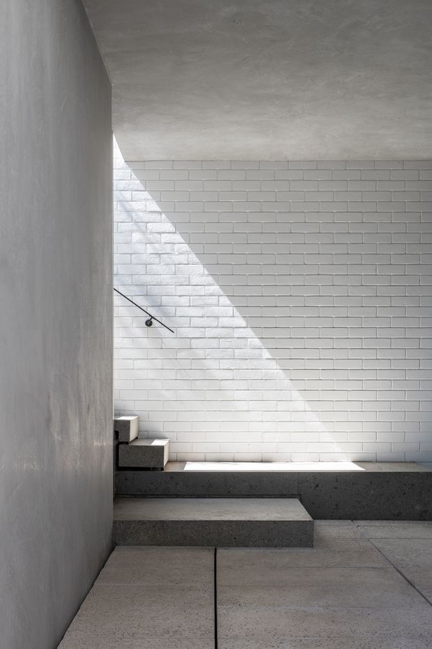 Minimalist interior stairway or landing with a dramatic triangular shaft of sunlight across a white painted brick wall. The space features raw concrete surfaces, a dark concrete stepped bench or stair edge, pale large-format floor slabs, and a simple black wall-mounted handrail. The composition is stark, geometric, and monochromatic.