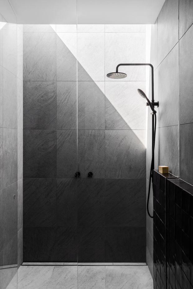 A narrow, minimalist walk-in shower lined with large-format grey stone-look tiles. A frameless glass screen separates the shower area, with a matte black overhead rain shower and handheld shower mounted above a dark tiled half-height partition. Strong diagonal sunlight creates a dramatic light-and-shadow pattern across the tiled wall and floor.