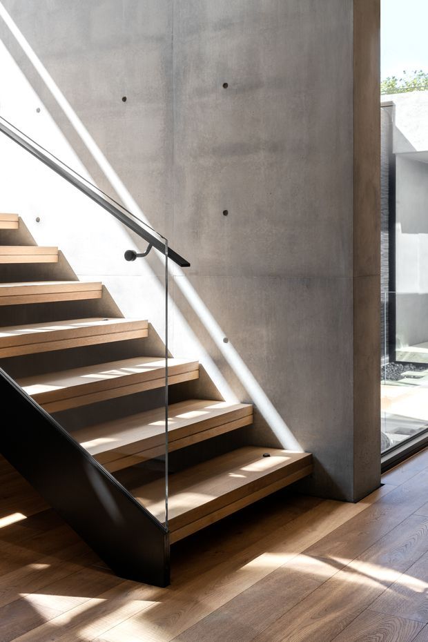 A contemporary interior stairway with floating timber treads supported by a dark steel structure and enclosed by a clear glass balustrade. The stair rises beside a tall smooth concrete wall, with warm timber flooring and strong natural sunlight creating diagonal shadows. Large glazing opens views toward a light-filled courtyard or exterior area.