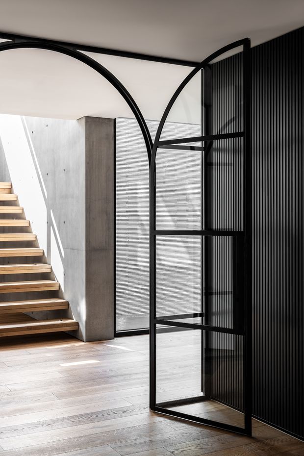A contemporary interior stair hall with floating timber stairs, pale timber flooring, a raw concrete wall, and a dramatic black metal and glass partition or pivot door. The partition features tall arched framing and vertical black slats, while full-height glazing with horizontal blinds brings in strong natural light.