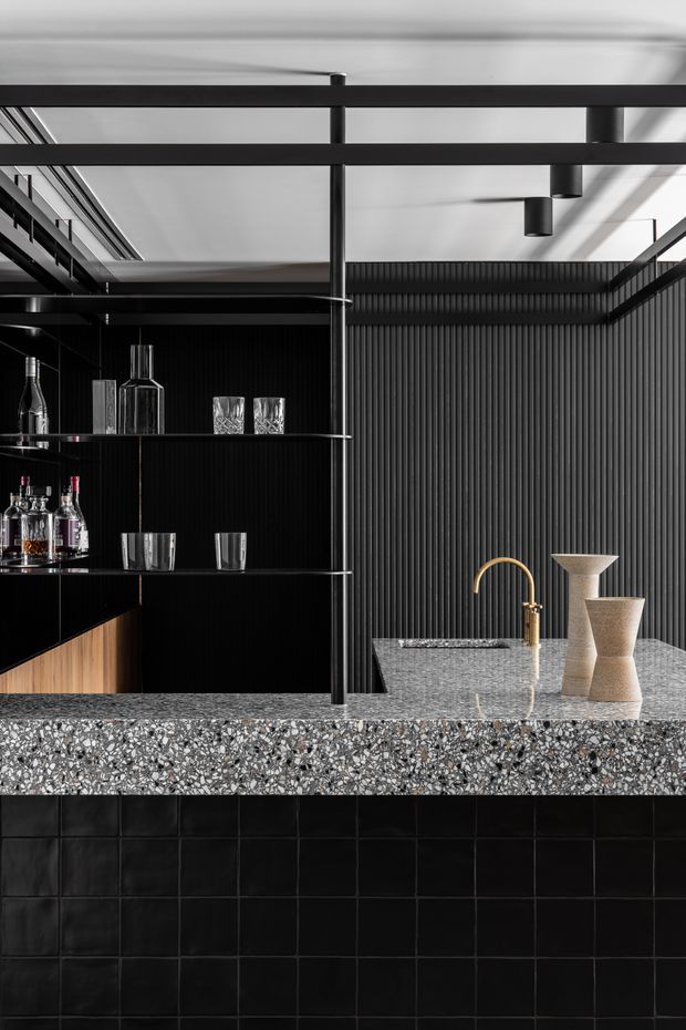 A dramatic, monochromatic home bar with a black vertically ribbed wall, open black metal shelving displaying bottles and glassware, and a substantial terrazzo island benchtop. The island has a dark tiled base and a curved brass tap, with sculptural neutral-toned vessels on the counter. Black steel framing and exposed linear ceiling elements reinforce the refined industrial character.