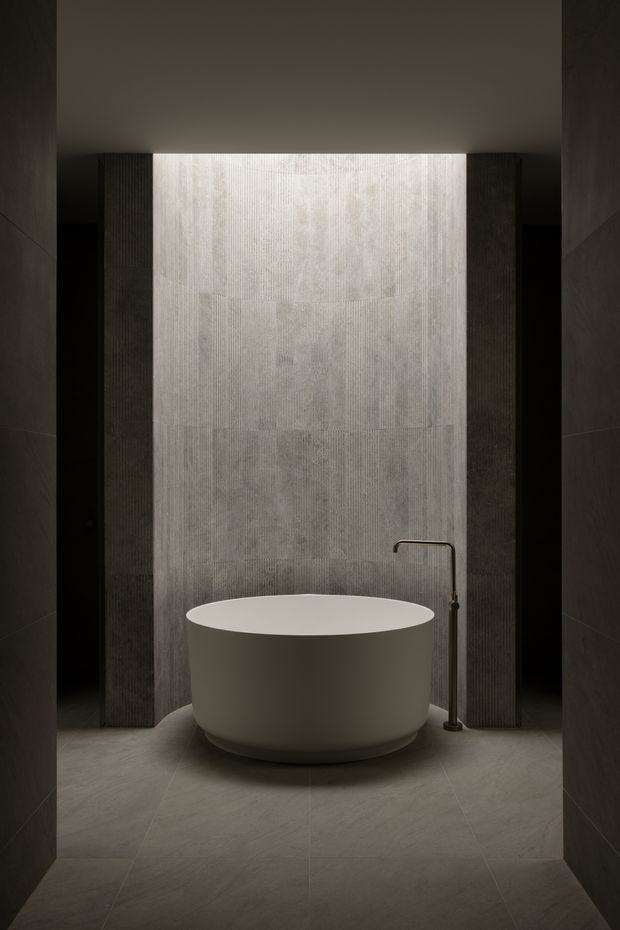 A moody, minimalist bathroom featuring a freestanding oval bathtub centered against a vertically textured grey feature wall. Dark vertical wall panels frame the bath, with a slim floor-mounted dark metal bath filler beside it. Large-format grey floor and wall tiles create a monolithic, refined composition with concealed lighting above the feature wall.