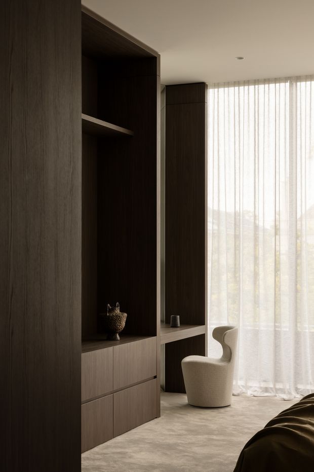 A softly lit contemporary bedroom with dark timber built-in cabinetry and open shelving, a compact integrated desk, and a sculptural upholstered armchair beside a full-height window. Sheer floor-to-ceiling curtains diffuse daylight, while a portion of the bed is visible in the foreground. The room has a warm, refined, minimalist feel with plush carpet and dark wood joinery.