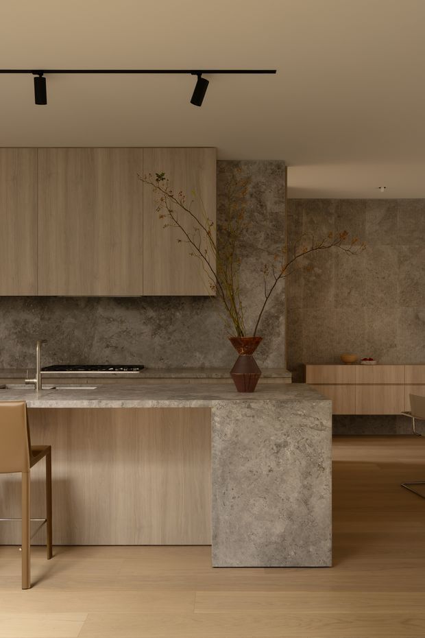 Warm, minimalist kitchen interior with a large light timber island and matching overhead cabinetry, grey natural-stone benchtops and splashback, integrated cooktop, and a substantial stone-clad island end panel. A sculptural vase with branches provides a restrained organic accent. The space opens toward a timber-floored dining or living area, with black linear track lighting and softly textured neutral wall finishes.