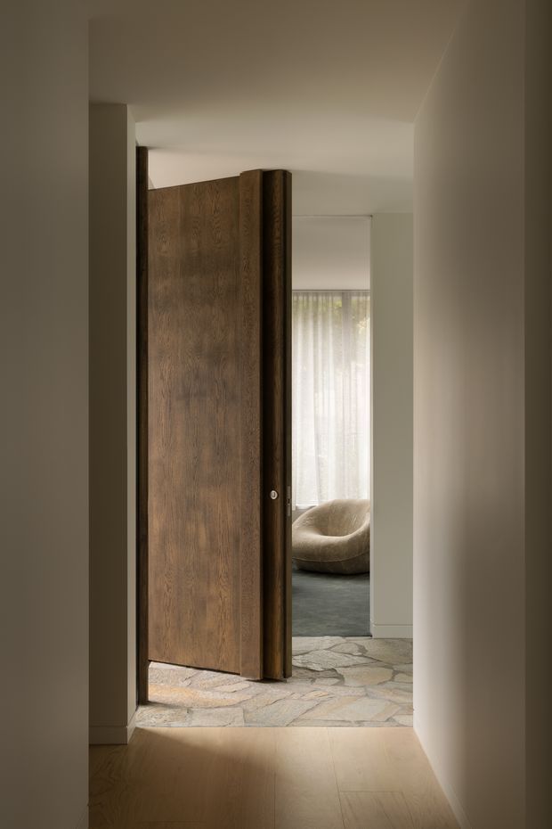 A warm, minimalist interior view from a narrow hallway through a partially open oversized timber pivot door. The doorway leads into a softly lit sitting or living area with sheer curtains, a rounded upholstered lounge chair, dark flooring, and an irregular natural-stone threshold or floor surface. Neutral plaster walls and pale timber flooring create a calm, refined atmosphere.