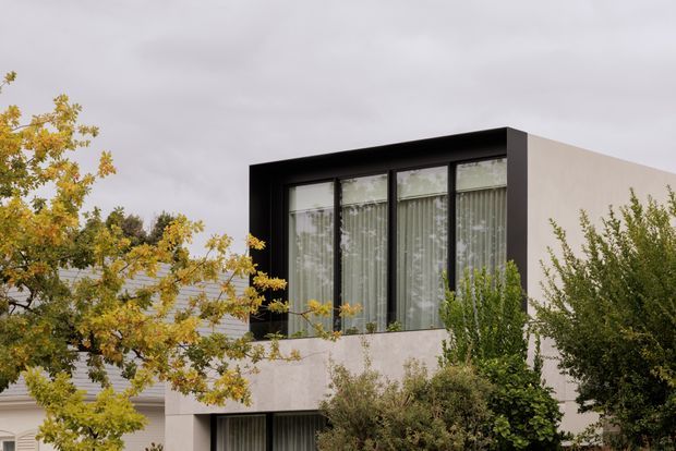 Exterior view of a contemporary two-storey residence, focused on a projecting upper-level window bay with a bold black frame and full-height glazing. Pale rendered walls, surrounding mature shrubs and autumn foliage, and an overcast sky create a restrained modern facade composition.