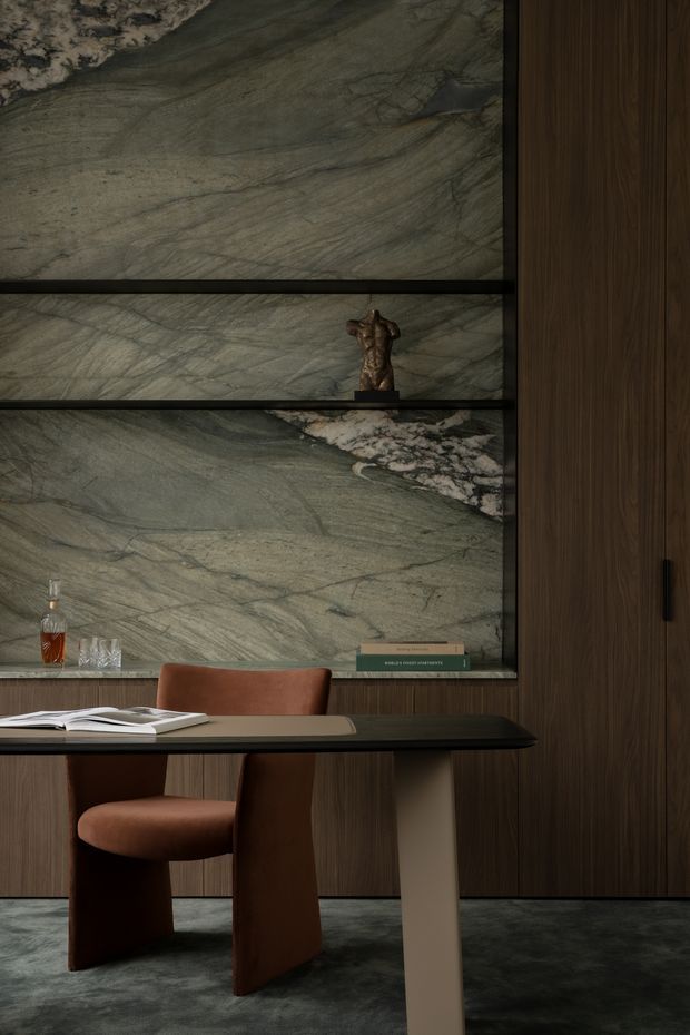 A moody, refined interior vignette featuring a dark timber built-in wall and full-height cabinetry, with a large veined green-grey natural stone feature wall and open display niches. A sculptural wood-toned desk with a dark top sits in front, paired with a curved rust-brown upholstered chair. Books, glassware, and a small sculpture add restrained decorative detail, while dark carpet grounds the space.