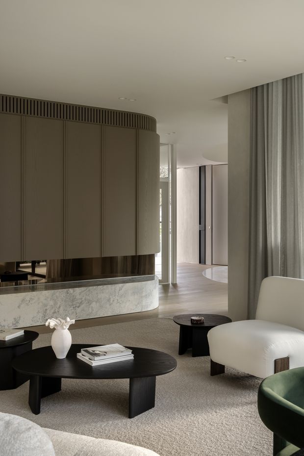 A refined contemporary open-plan living room with a softly curved built-in feature and stone-clad base, sculptural black oval coffee tables, rounded cream lounge chairs, textured carpet, and full-height grey curtains. A partially visible adjoining hallway and curved timber-toned flooring extend into the background. The palette is muted and neutral, with layered curves, concealed lighting, and carefully integrated joinery creating a calm, luxurious atmosphere.