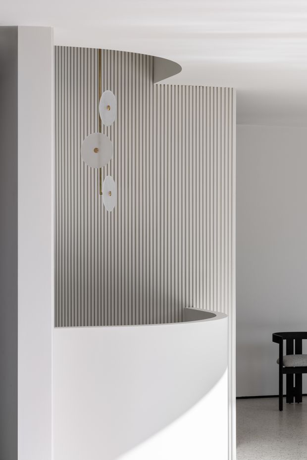 A minimalist interior featuring a sculptural curved white partition or stair balustrade with a vertically fluted wall behind it. Three simple circular white wall lights with small brass-toned centres decorate the fluted surface. A black dining chair is partially visible at right, and the space has a restrained white and grey palette with terrazzo-like flooring.