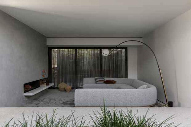 A refined contemporary living room with a large curved light-grey sectional sofa facing a minimalist linear fireplace and floating media shelf. Full-height dark sheer curtains cover the rear glazing, while a sculptural arched floor lamp adds a strong graphic element. The room uses a restrained palette of concrete grey, white, black, and soft grey furnishings, with ornamental grasses visible in the foreground.