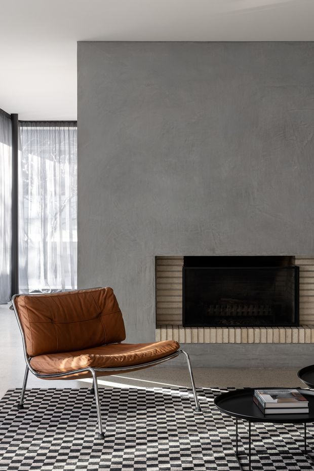 A refined living room centered on a large textured grey plaster fireplace wall with a black firebox and horizontal light brick hearth detailing. A cognac-brown leather sling chair with tubular metal legs sits over a bold black-and-white checkerboard rug, beside a low black round coffee table with books. Sheer floor-to-ceiling curtains bring in strong natural light, creating a modern, graphic interior.