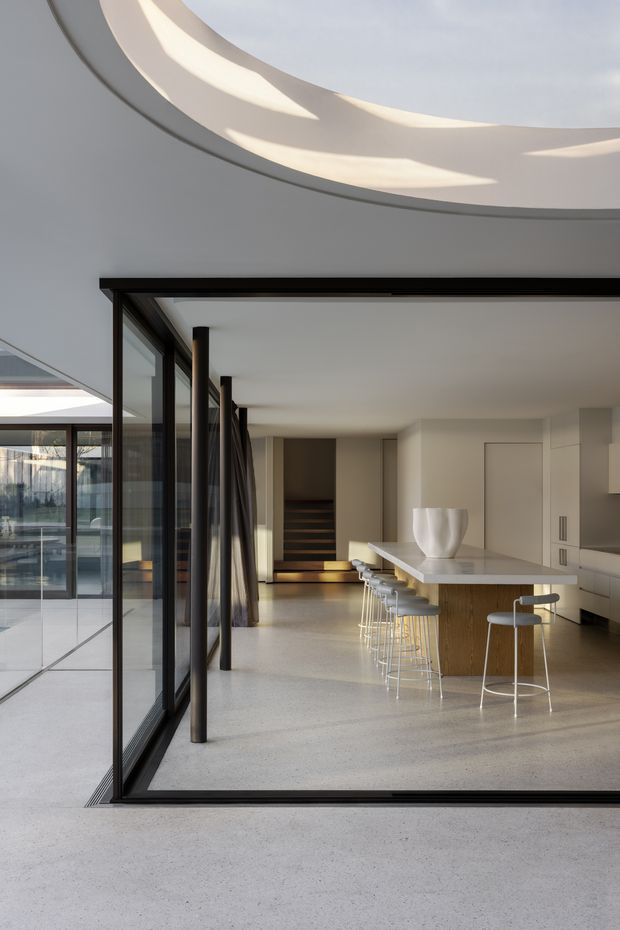 A refined contemporary open-plan kitchen and living area viewed through fully glazed black-framed sliding doors. The space features a long white island with integrated seating, pale built-in cabinetry, polished concrete flooring, and a sculptural white vessel. A large curved ceiling opening or skylight introduces soft daylight, while dark vertical framing and a stair-like timber feature add contrast.