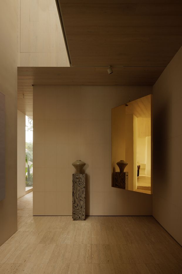 A warm, minimalist interior hallway or entry space with continuous pale timber wall and ceiling lining, matching light timber flooring, and a large reflective bronze-toned wall panel. A sculptural pedestal and vessel form a focal point, while an opening at left provides a glimpse of greenery outside. Recessed and track-style lighting create a calm, gallery-like atmosphere.