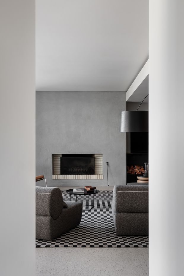 A refined contemporary living room viewed through a narrow opening, featuring a grey textured feature wall with a linear fireplace, two rounded grey upholstered lounge chairs, a black-and-white checkerboard rug, a low dark coffee table, and a large black pendant light. Dark built-in shelving with stacked firewood is visible to the right. The palette is monochromatic, with clean architectural lines and a sophisticated, moody atmosphere.
