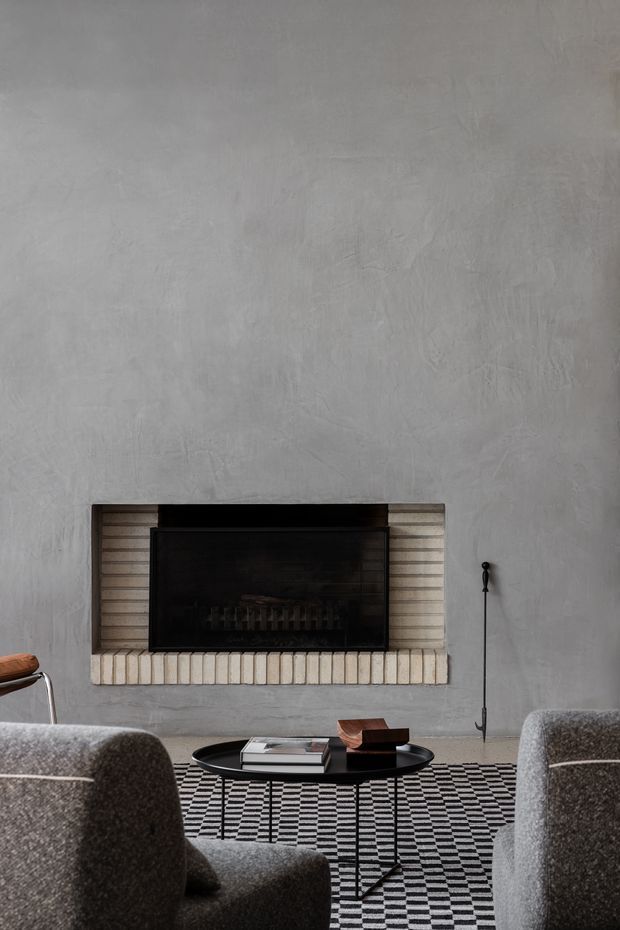 A moody contemporary living room centered on a recessed black fireplace set into a textured grey plaster wall with light brick surround. Two substantial grey upholstered lounge chairs frame a low black oval coffee table, while a bold black-and-white geometric rug adds graphic contrast. The space has a restrained, refined palette and minimalist furnishings.