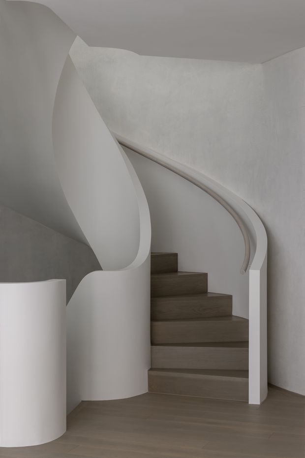 A sculptural interior stairway with a sweeping curved white balustrade and rounded wall forms. The staircase has broad dark timber-toned treads and risers, set against smooth white plaster walls and a muted neutral floor. The design is minimalist and highly sculptural, with soft organic curves and subdued natural tones.