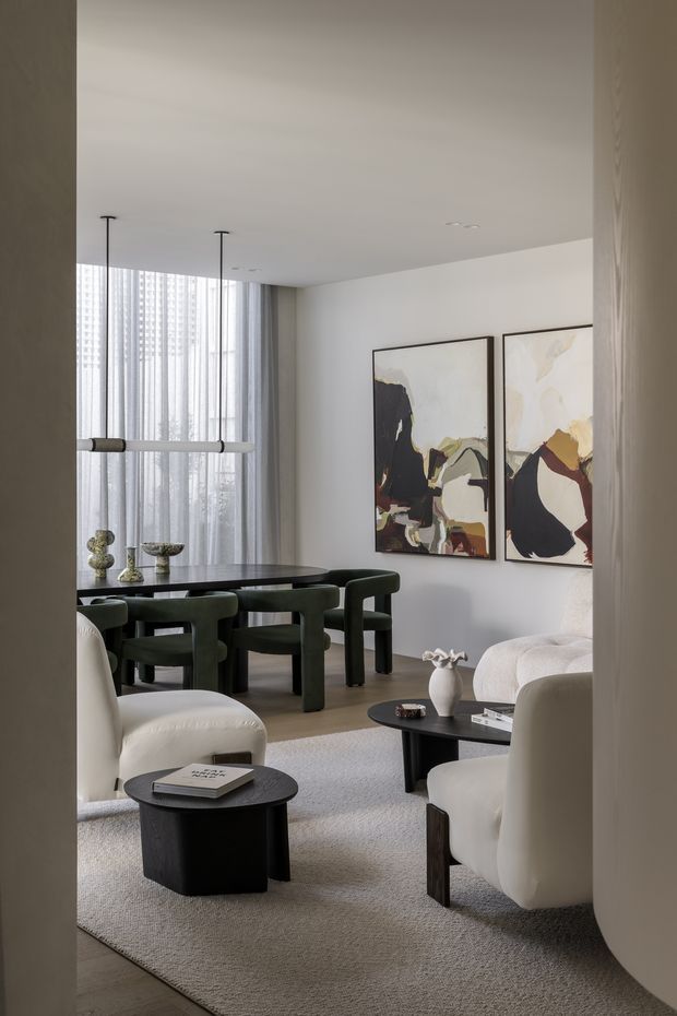 A refined open-plan living and dining area viewed through a doorway. The room features a dark oval dining table with deep green upholstered chairs, sculptural white lounge chairs, black coffee tables, a large light neutral rug, abstract artwork in dark frames, sheer floor-to-ceiling curtains, and slim linear pendant lighting. The palette is restrained in white, grey, black, and forest green, creating a calm contemporary atmosphere.