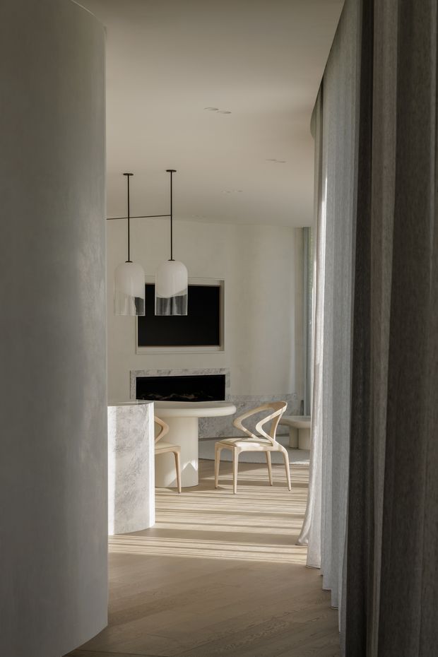 A softly lit minimalist interior viewed through a curved opening or corridor, leading into an elegant dining and living area. The space features pale timber flooring, sculptural light-coloured dining chairs, a rounded dining table, a marble or stone-clad island/console, a dark built-in fireplace or media niche, pendant lights with opal glass shades, and full-height sheer curtains. The palette is warm and restrained, with curved architectural forms and refined contemporary detailing.