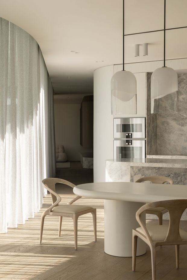 A refined open-plan dining and kitchen interior in a soft neutral palette. A rounded cream dining table is surrounded by sculptural timber-framed chairs, positioned beside full-height sheer curtains that cast linear sunlight across the pale timber floor. The kitchen features curved white cabinetry, integrated stainless-steel ovens, a pale veined stone island and splashback, and two oversized translucent pendant lights. A darker living area is visible beyond the kitchen.