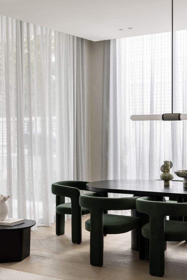 A refined contemporary dining area with a round dark dining table and sculptural deep-green upholstered dining chairs. Full-height sheer curtains wrap the corner windows, creating a bright, softly filtered setting. The room features pale timber flooring, warm light-grey walls, a simple white ceiling with recessed downlights, and a minimalist suspended linear pendant. Decorative ceramic objects sit on the table, while a small dark side table is visible in the foreground.