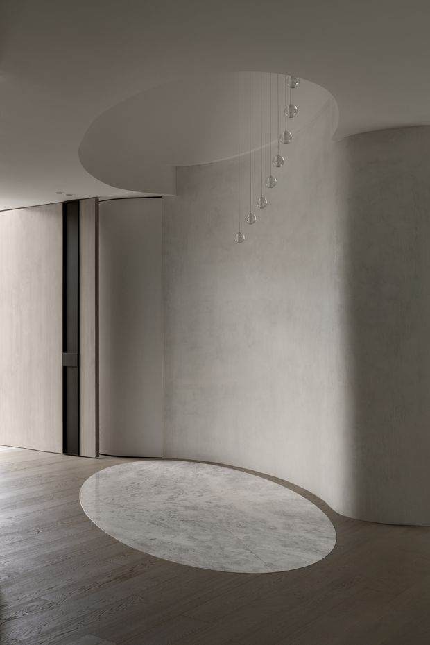 A refined minimalist interior featuring a curved plastered wall and sculptural curved ceiling soffit, dark timber flooring, a pale oval stone or terrazzo-like inset floor feature, and a delicate cluster of suspended glass pendant lights. The palette is monochromatic and warm grey, with concealed or integrated door detailing and a calm, gallery-like atmosphere.