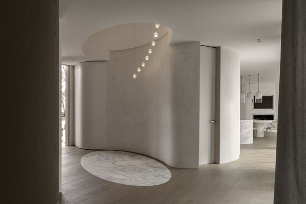 A refined minimalist interior viewed through a dark-framed opening, featuring a sculptural curved plaster partition and rounded ceiling soffit with a cascading line of small pendant lights. Pale timber flooring, soft neutral surfaces, and a circular rug create a calm entry or circulation space, with a dining and kitchen area visible beyond.
