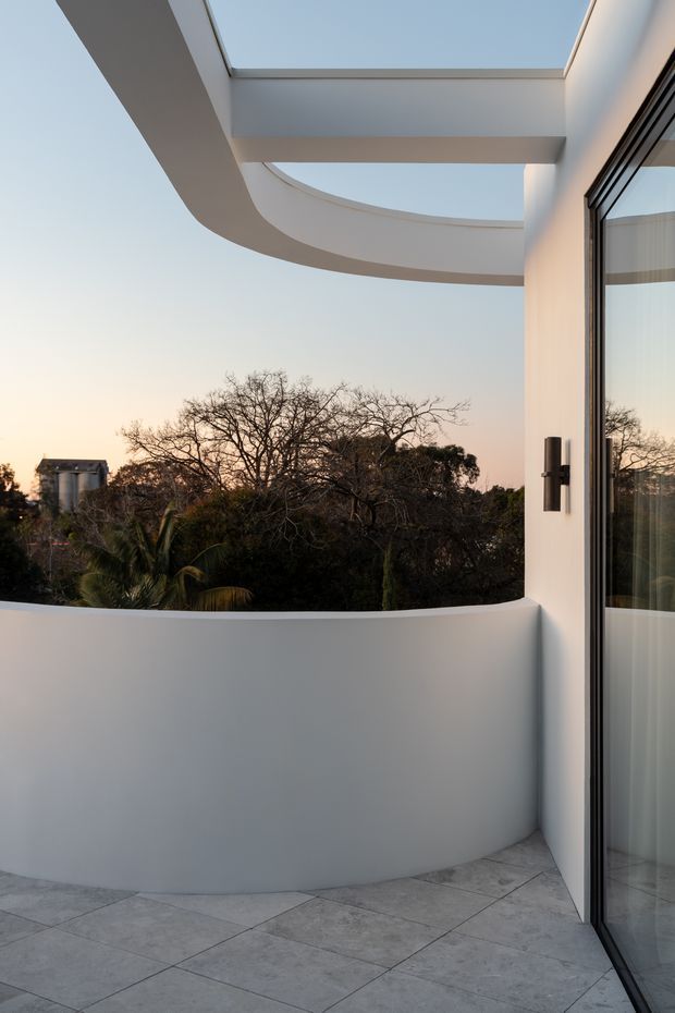 A curved white architectural terrace or balcony with a low parapet, pale tiled flooring, and a large black-framed glazed door. The space is defined by sculptural curved beams and an overhead skylight/opening, overlooking mature trees at sunset. The design is clean, minimalist, and strongly contemporary.