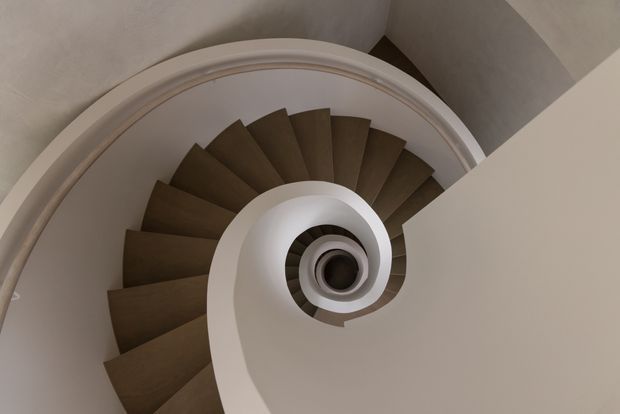 Overhead view looking down a dramatic spiral staircase, with dark timber or wood-toned wedge-shaped treads wrapping around a central opening and a smooth white curved balustrade forming a strong spiral. The surrounding walls and ceiling are warm neutral plaster, creating a sculptural, refined architectural composition.