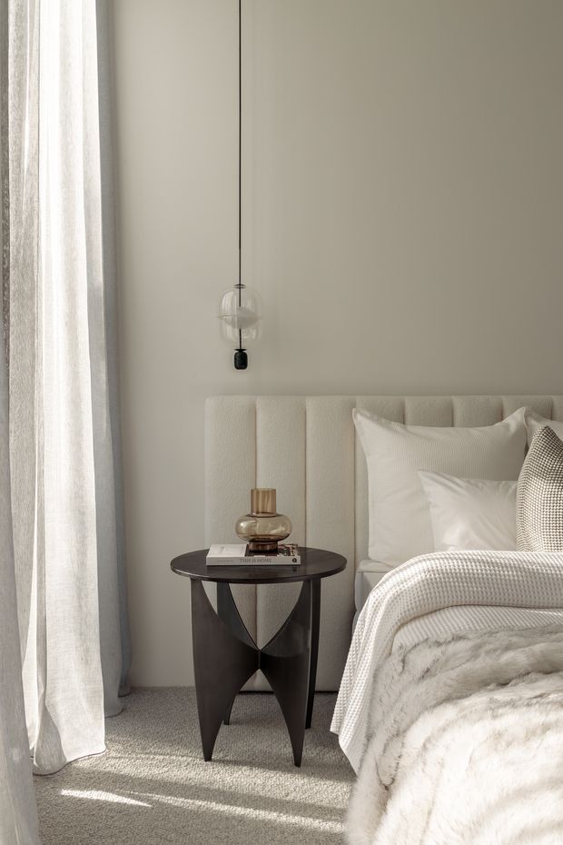 A softly lit bedroom corner featuring a plush upholstered bed with a tall vertically channelled cream headboard, layered white and neutral bedding, a dark round bedside table with sculptural legs, and a delicate pendant light. Floor-to-ceiling sheer curtains filter daylight beside a pale neutral wall, while textured carpet and a folded faux-fur throw add softness.