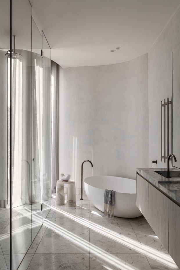 A bright, refined contemporary bathroom featuring a freestanding white bathtub, frameless glass shower enclosure, double vanity with stone benchtop, wall-mounted dark metal towel warmer, and large light marble-look floor tiles. Soft sheer curtains and strong sunlight create a calm, luxurious atmosphere.