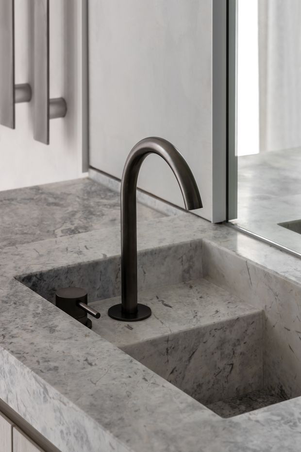 Close-up of a minimalist bathroom vanity featuring a sculptural dark metal arched tap and separate cylindrical handle set into a grey-veined natural stone benchtop with an integrated stone basin. Pale fixed cabinetry and a frameless glass shower or partition are visible in the background.