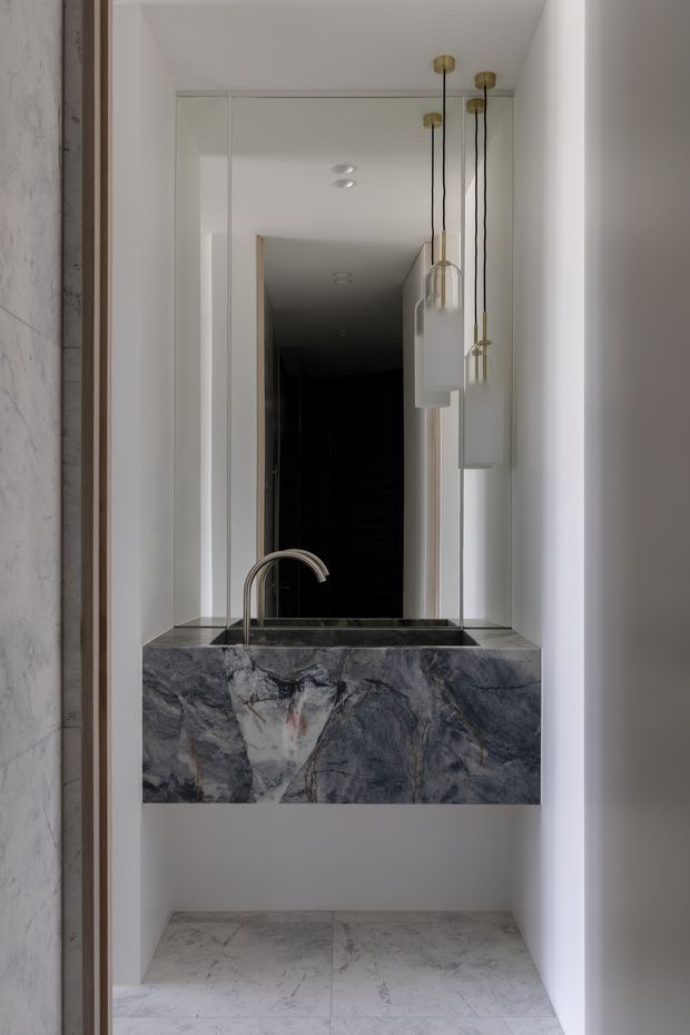 A compact, refined washroom vanity viewed straight on. A dramatic dark veined stone basin and matching waterfall-front counter sit beneath a large mirror, with a curved brushed-metal tap and three suspended glass pendant lights. White walls and pale veined floor tiles create a high-contrast, gallery-like composition, while a dark tiled passage is reflected behind the vanity.