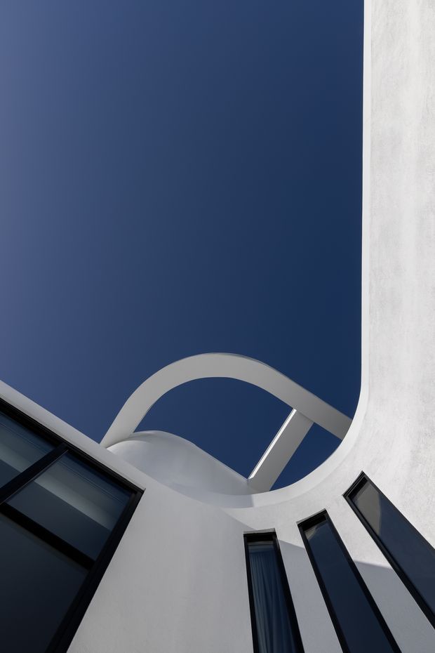 Upward-looking architectural view of a sculptural white curved facade or courtyard, featuring a large circular/arched opening framing the deep blue sky and tall, narrow dark-framed windows around the perimeter. The composition emphasizes geometric curves, strong contrast, and modern minimalist architecture.