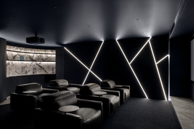 A dark, high-end home cinema with rows of plush black leather recliner seating, a black carpeted floor, and dramatic geometric linear LED lighting across the black feature wall. Illuminated marble-effect display shelving and a ceiling-mounted projector are visible at the rear, creating a luxurious, cinematic atmosphere.