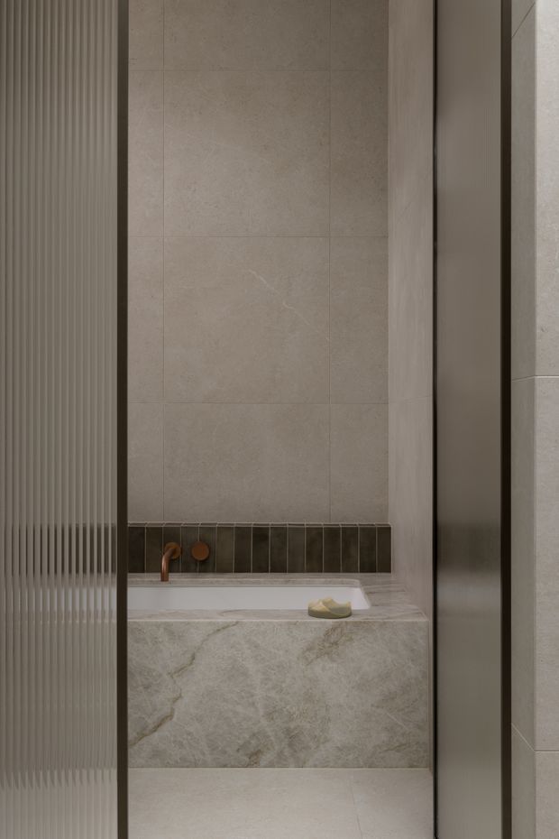 A close-up view of a refined contemporary bathroom, focused on a built-in bathtub beneath large-format warm grey stone-look wall tiles. The tub surround and benchtop feature strongly veined pale natural or engineered stone, with a slim dark vertical tile backsplash and bronze-toned wall-mounted tapware. A fluted translucent glass panel frames the left side, while dark slim framing and neutral finishes create a calm, understated atmosphere.