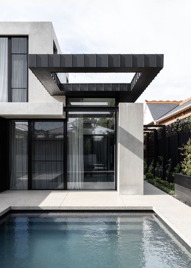 Contemporary multi-level residential exterior viewed from a swimming pool courtyard. The facade combines smooth pale concrete or rendered walls, dark aluminium-framed floor-to-ceiling glazing, and a prominent projecting black slatted canopy or bridge connecting the upper levels. A narrow planted side walkway and neighbouring pitched-roof structures are visible, with the pool occupying the foreground.