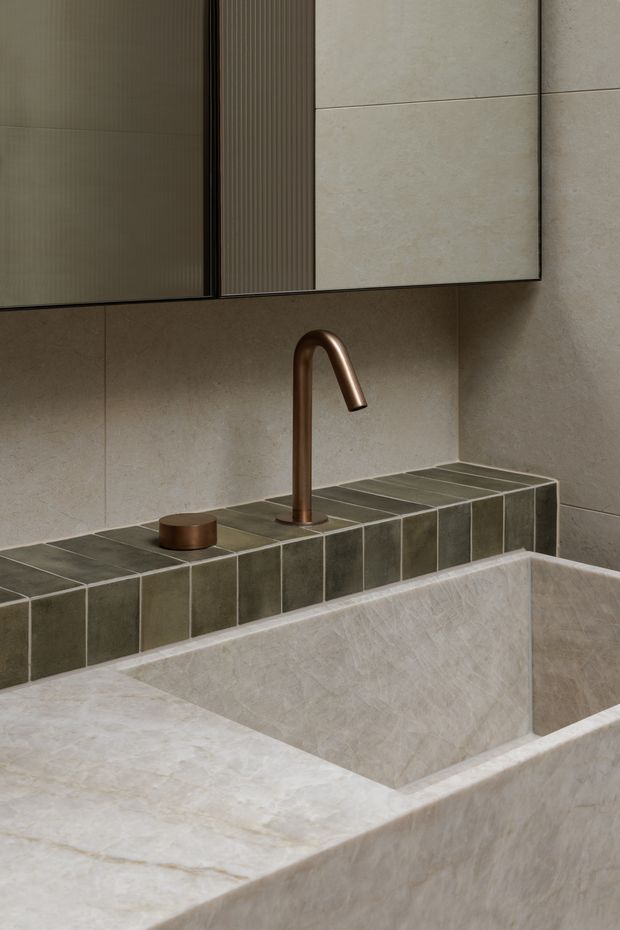 Close-up of a refined bathroom vanity featuring a large integrated pale stone basin, muted olive-grey square tiled splashback and ledge, a brushed bronze gooseneck tap and matching control, and wall-mounted mirrored cabinetry with ribbed detailing. The materials and restrained palette create a warm, minimalist, high-end atmosphere.