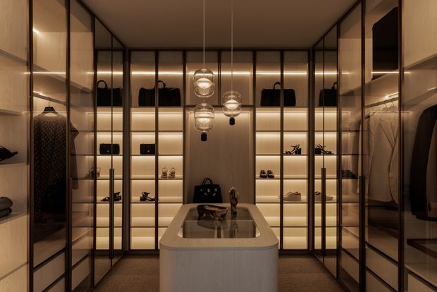 A luxurious walk-in wardrobe or dressing room with floor-to-ceiling glazed cabinetry, warm integrated LED lighting, dark-framed glass doors, and illuminated shelving displaying handbags, shoes, and accessories. A central island with rounded corners and a reflective inset top anchors the room, while pendant lights create a refined, intimate atmosphere.