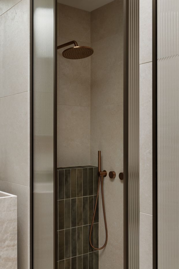 Close-up view of a refined walk-in shower enclosure. Large warm beige stone-look wall tiles surround a darker vertically tiled lower wall section, with a bronze/copper-toned round rainfall showerhead, handheld shower, and controls. Ribbed translucent glass panels and dark slim frames define the enclosure, creating a warm, subdued contemporary bathroom aesthetic.