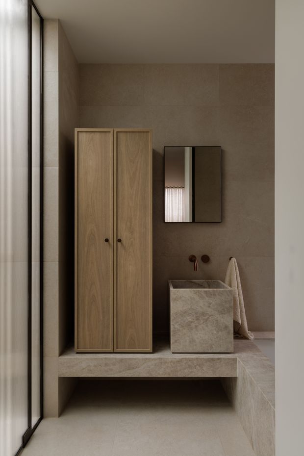 A compact, refined bathroom with warm beige stone slab walls and flooring, a floating stone vanity with integrated basin, timber cabinetry, a black-framed mirrored cabinet, and brushed copper wall-mounted tapware. Full-height glazing with a sheer blind brings in soft natural light, creating a calm, minimalist atmosphere.