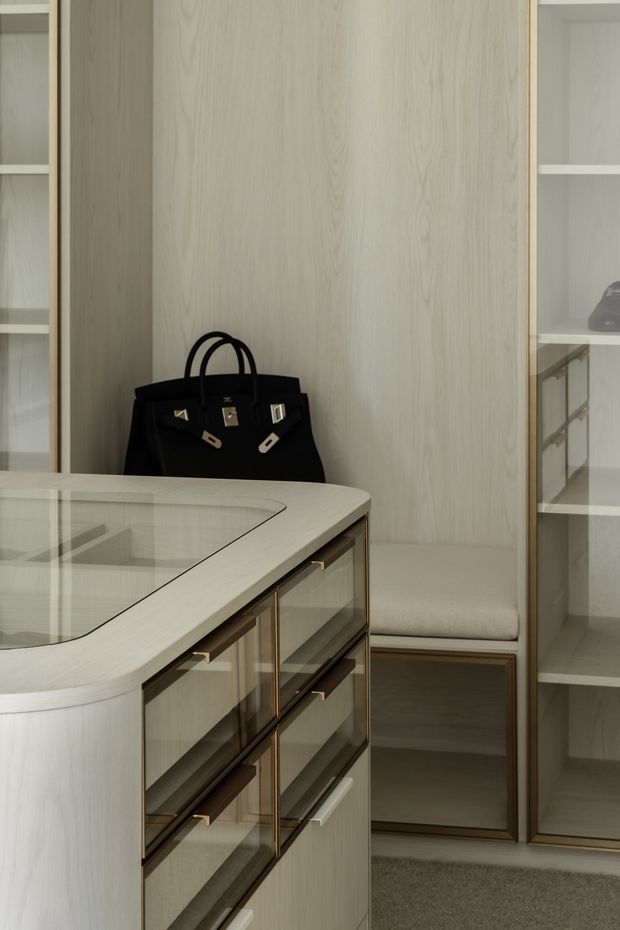 A refined walk-in wardrobe or dressing room with pale timber wall panelling, open shelving, built-in drawers, glass-fronted cabinetry, and a curved central island display cabinet. Brass-toned trim and handles add a subtle luxury accent, while a black handbag and neutral carpet provide contrast. The space has a calm, softly lit, bespoke built-in design.