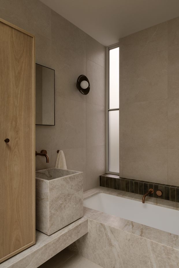 A warm, minimalist bathroom clad in large beige stone-look tiles, with a freestanding stone-clad basin, integrated bathtub, dark tiled ledge, timber door, slim vertical window, wall-mounted mirror, and dark bronze fixtures.
