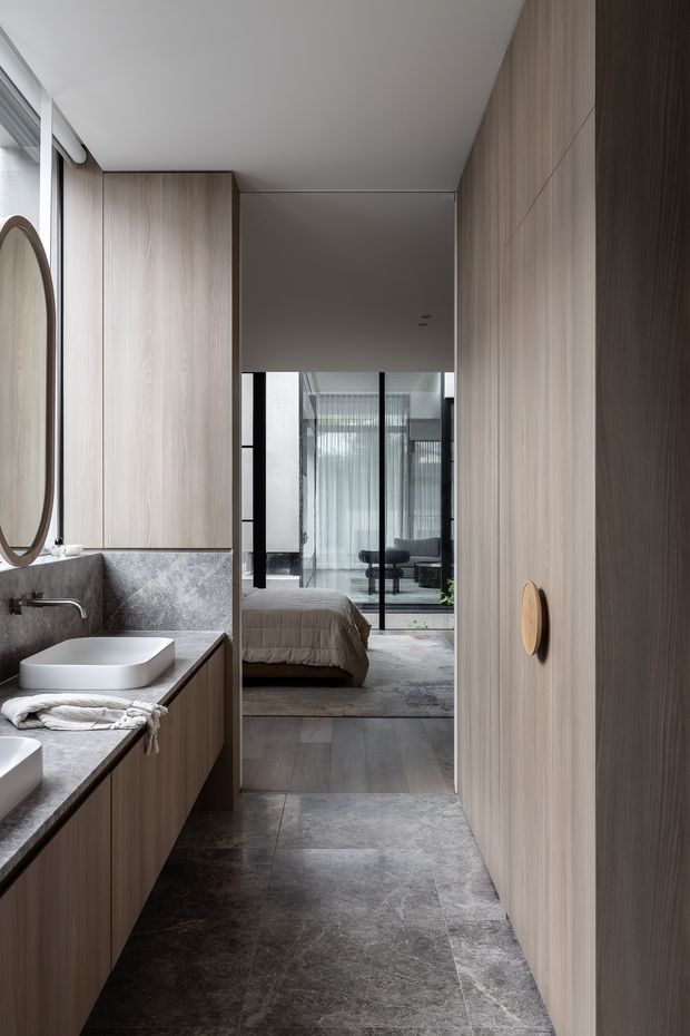 A refined contemporary bathroom corridor with a long floating timber vanity, dark stone countertop and backsplash, white vessel basins, wall-mounted chrome tapware, and a large round mirror. Full-height light timber cabinetry lines both sides, leading directly into a bedroom visible through black-framed glazing. Large-format dark stone-look floor tiles unify the space, while sheer curtains and restrained furnishings create a calm, minimalist atmosphere.