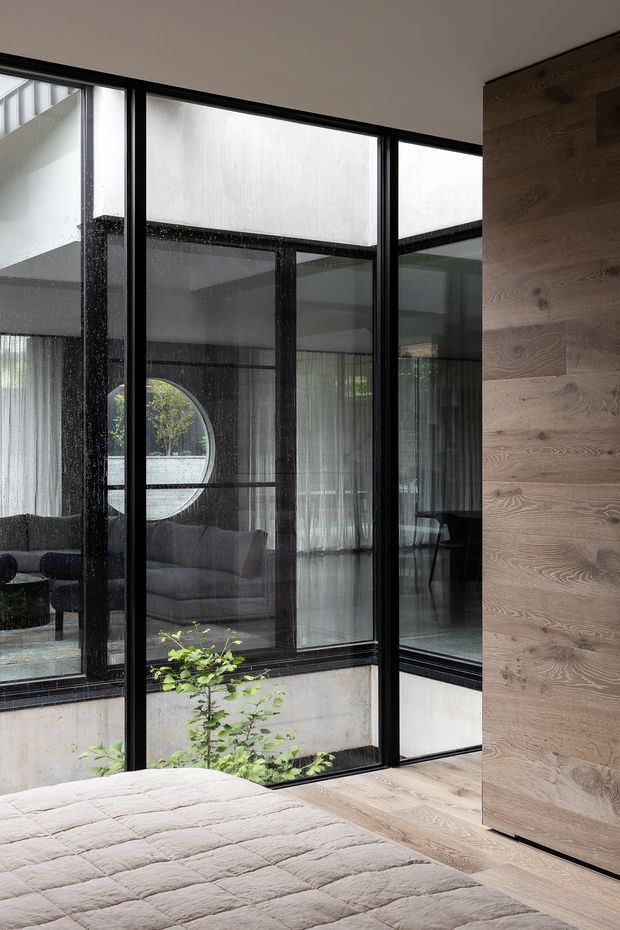 A contemporary bedroom with floor-to-ceiling black-framed glazing overlooking an internal courtyard. The room features a pale quilted bedspread, warm oak wall panelling and flooring, and a reflective view into an adjacent lounge with a curved window and grey sofa. The glazing, courtyard planting and dark structural framing create a refined indoor-outdoor connection.