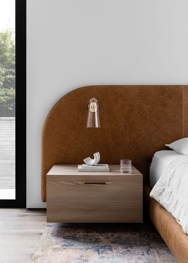 Close-up interior view of a refined bedroom corner featuring a warm cognac-brown upholstered bed with a large rounded headboard, a wood bedside cabinet with a slim brass and leather pull, a wall-mounted reading light, and layered neutral bedding. A textured patterned rug sits over pale timber flooring, while a full-height black-framed glazed opening brings in greenery and daylight.