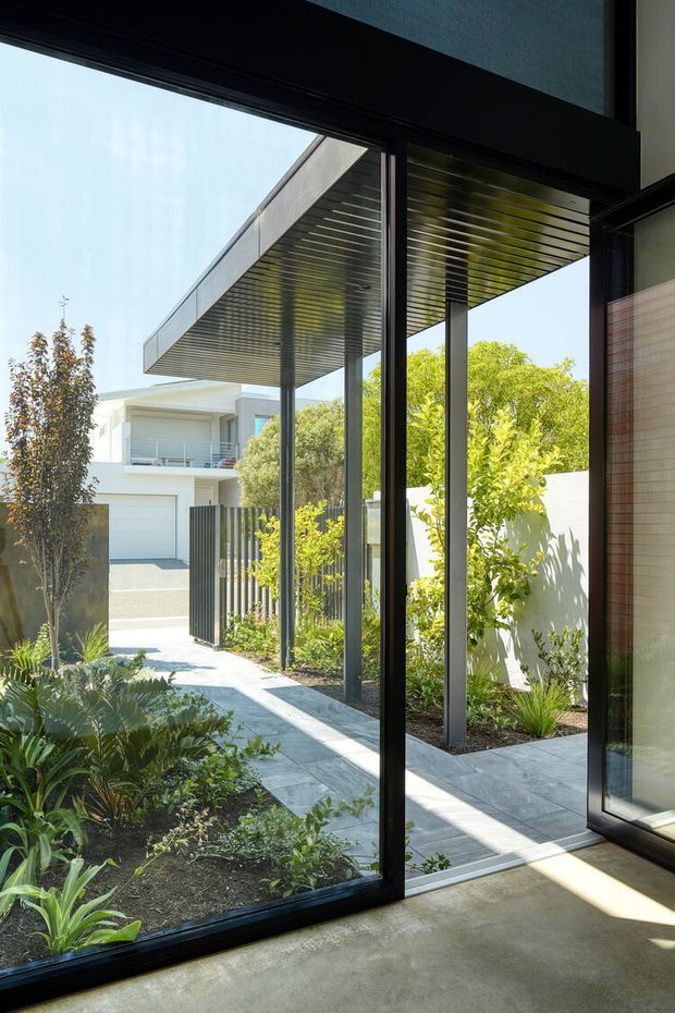 View from inside a contemporary home through large black-framed glazed doors toward a landscaped courtyard and paved entry path. A deep projecting flat canopy with exposed linear soffit spans slender dark posts, while a neighbouring white modern residence, boundary fencing, garden beds, and mature planting are visible beyond. The interior foreground has a smooth polished concrete floor and a warm-toned wall surface.