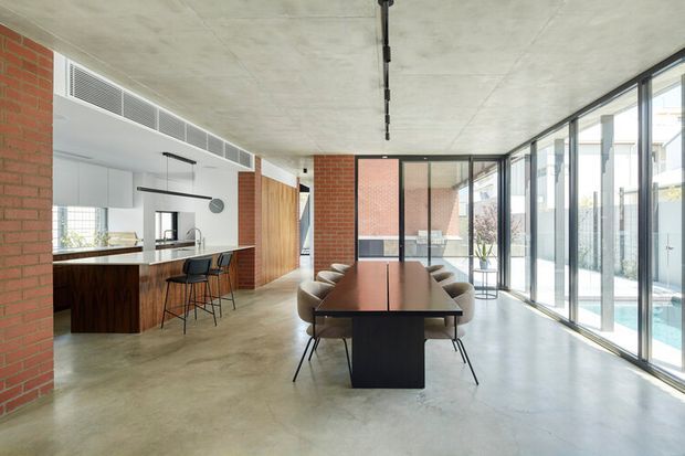 Open-plan contemporary interior combining a kitchen, dining area and living space. Exposed red-brick structural walls, a polished concrete floor and ceiling, dark timber kitchen cabinetry, and extensive full-height glazing define the space. A long dining table with upholstered chairs sits centrally, while a compact kitchen island with bar stools occupies the left side. The glazing opens views toward a narrow pool or water feature and adjacent urban buildings.